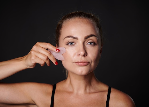 Foto de estudio de una bonita mujer caucásica haciendo un masaje facial de drenaje linfático con un masajeador de guasha de piedra de jade Tratamiento de spa de belleza Primer plano Tratamiento de belleza de rejuvenecimiento suavizante antienvejecimiento