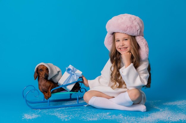 foto de estudio de una chica con un suéter blanco y un sombrero y un perro salchicha sentado en un trineo