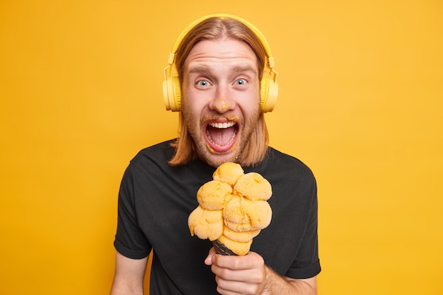 Foto de estudio de chico pelirrojo emocional sucio después de comer helado de cono disfruta de un delicioso postre de verano se esfuerza por la comida sabrosa escucha la pista de audio en los auriculares