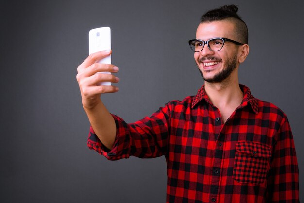 Foto de estudio de guapo turco con anteojos mientras usa el teléfono móvil contra el fondo gris