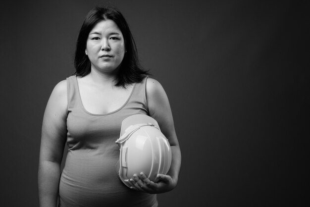 Foto de estudio de hermosa mujer asiática con sobrepeso, trabajador de la construcción con casco contra un fondo gris en blanco y negro