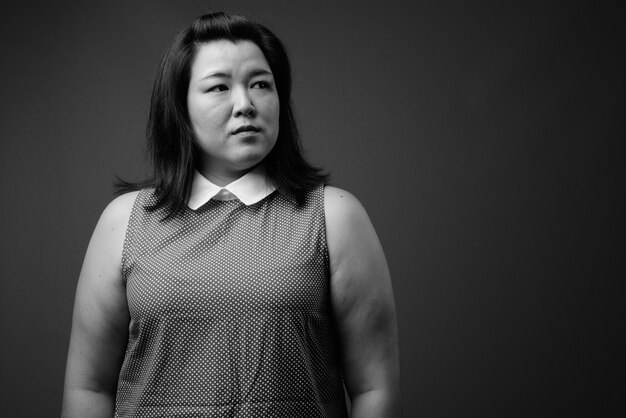 Foto de estudio de hermosa mujer asiática con sobrepeso vistiendo un vestido contra un fondo gris en blanco y negro