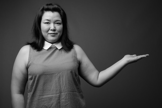 Foto de estudio de hermosa mujer asiática con sobrepeso vistiendo un vestido contra un fondo gris en blanco y negro