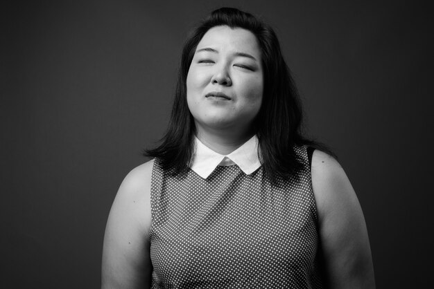 Foto de estudio de hermosa mujer asiática con sobrepeso vistiendo un vestido contra un fondo gris en blanco y negro