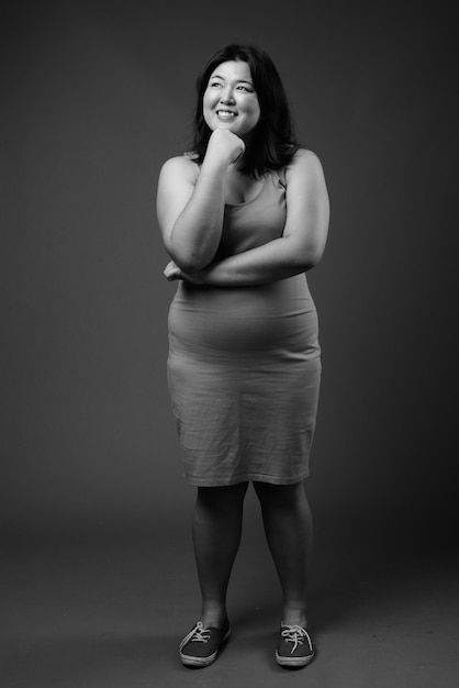 Foto de estudio de hermosa mujer asiática con sobrepeso vistiendo un vestido sin mangas contra un fondo gris en blanco y negro