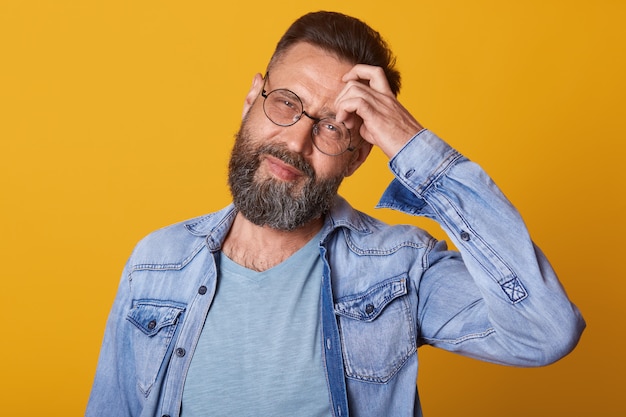 Foto de estudio de hombre guapo con barba toca su frente y tiene dolor de cabeza, vestido de mezclilla y camiseta gris