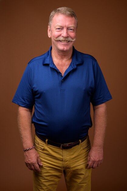 Foto de estudio de hombre senior guapo con bigote vistiendo camisa polo azul contra marrón