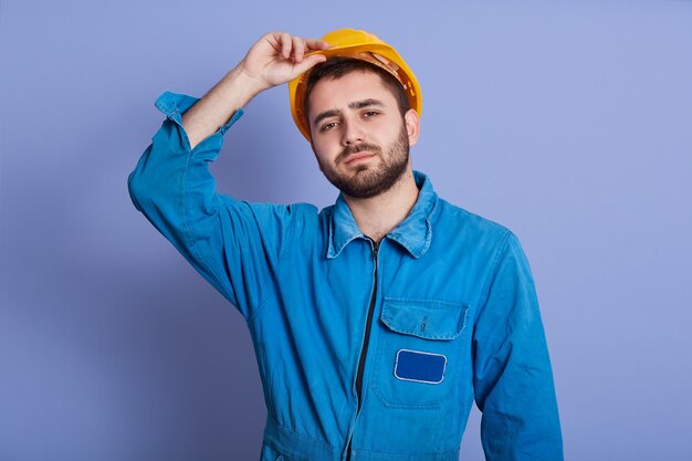 Foto foto de estudio del hombre vestido con uniforme azul y casco de construcción amarillo mirando a la cámara, manteniendo la mano en su casco, chico guapo con barba oscura, parece cansado después del trabajo duro. concepto de construcción.