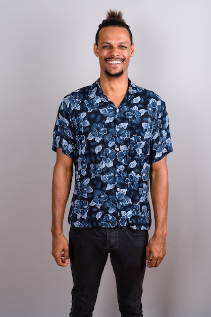 Foto de estudio de joven apuesto hombre africano barbudo vistiendo camisa hawaiana con el pelo atado contra un fondo gris