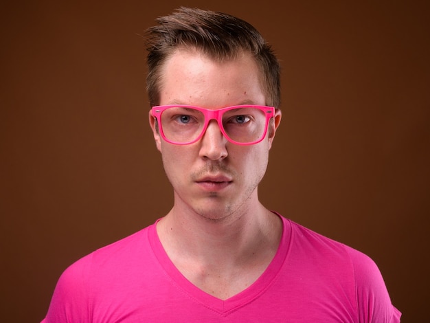 Foto de estudio de joven apuesto hombre vestido con camisa rosa con anteojos rosa a juego contra el fondo marrón