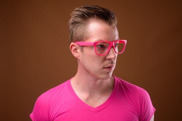 Foto de estudio de joven apuesto hombre vestido con camisa rosa con anteojos rosa a juego contra el fondo marrón