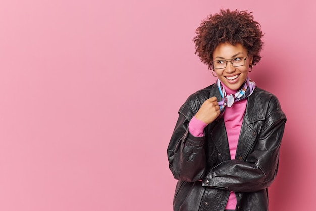 Una foto de estudio de una joven de cabello bastante rizado sonríe felizmente mira hacia otro lado usa una chaqueta de cuero negro y un pañuelo alrededor del cuello tiene una expresión alegre aislada sobre un espacio de copia en blanco de fondo rosa