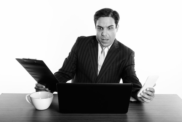 Foto de estudio del joven empresario persa sentado detrás de un escritorio de madera aislado, blanco y negro