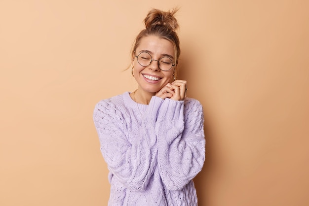 La foto de estudio de una joven feliz mantiene las manos cerca de la cara sonríe con dientes tiene una expresión facial tierna y romántica recuerda algo agradable en mente usa anteojos redondos y suéter de punto cálido