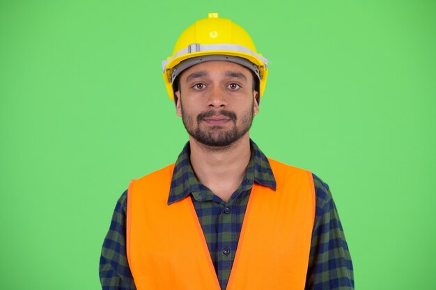 Foto de estudio de joven guapo persa barbudo trabajador de la construcción contra chroma key con fondo verde