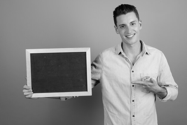 Foto de estudio de joven guapo sosteniendo pizarra contra un fondo gris en blanco y negro