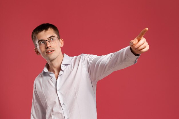 Foto de estudio de un joven inteligente con una camisa blanca clásica, relojes negros y anteojos apuntando a algo mientras posa sobre un fondo rosa. Corte de pelo con estilo. Concepto de emociones sinceras. Copie el espacio.