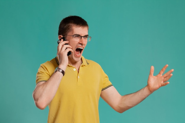 Foto de estudio de un joven inteligente con una camiseta informal amarilla, anteojos y relojes negros hablando por teléfono y gritando a alguien mientras posa sobre un fondo azul. corte de pelo con estilo. emo sincero