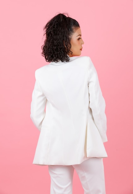 Una foto de estudio de una joven y sexy mujer de negocios profesional asiática con un peinado rizado y un traje casual de moda blanco de pie posando cogidos de la mano en los bolsillos de los pantalones volviendo a un fondo rosa