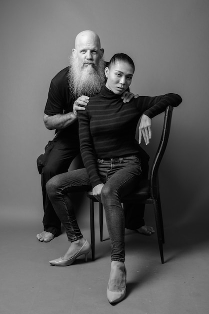 Foto de estudio de maduro hombre calvo barbudo y joven y bella mujer asiática juntos contra la pared gris en blanco y negro