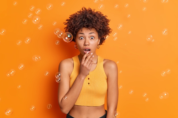 Foto de estudio de mujer afroamericana sorprendida se queda sin palabras mantiene la boca abierta de maravilla vestida con top recortado reacciona ante noticias asombrosas aisladas sobre pared naranja burbujas de jabón alrededor