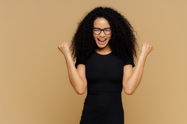 Foto de estudio de una mujer alegre y alegre con el pelo rizado, levanta los puños cerrados