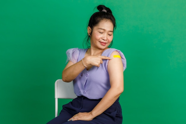 Foto de estudio de una mujer asiática de mediana edad sentada con una sonrisa apuntando al vendaje de yeso amarillo en el brazo después de recibir la vacunación contra el coronavirus covid 19 por parte del médico en la clínica sobre fondo verde.