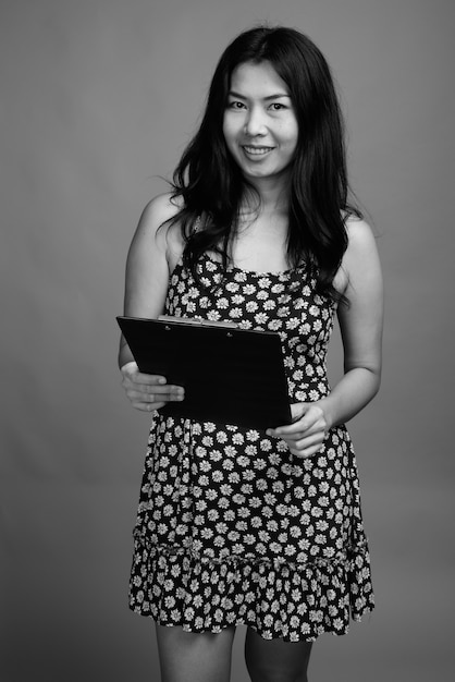 Foto de estudio de mujer asiática vistiendo un vestido mientras sostiene el portapapeles contra un fondo gris en blanco y negro