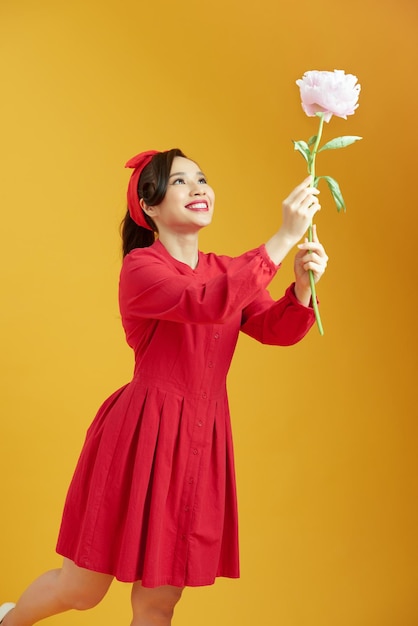 Una foto de estudio de una mujer encantadora aislada sobre un fondo de estudio amarillo sosteniendo una flor de peonía blanca con una expresión complacida