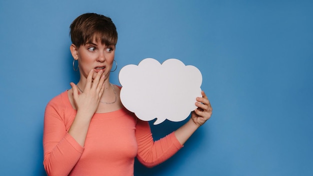 Foto de estudio de una mujer joven con una expresión perpleja que sostiene una pancarta blanca vacía en forma de nube sobre un fondo azul. Copia espacio para tu anuncio o texto.