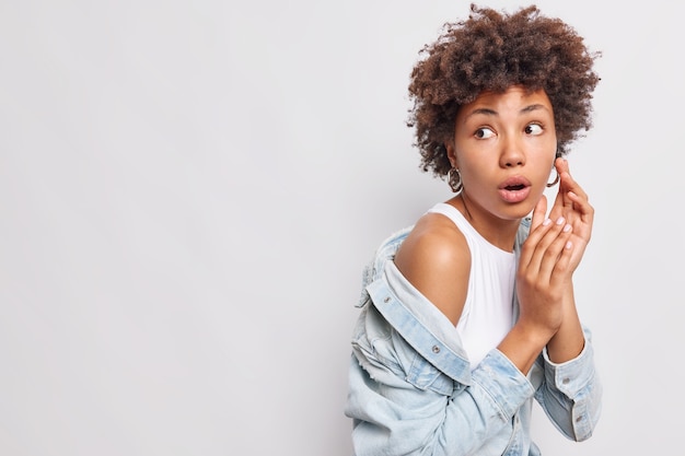 Foto de estudio de una mujer de pelo rizado impresionado que parece asombrado mira fijamente con total incredulidad viste una camiseta blanca y una camisa de mezclilla aislada sobre una pared blanca