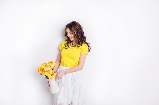 Una foto de estudio de una niña de pie y sosteniendo un jarrón blanco con flores. La niña tiene el cabello castaño largo y ondulado y viste una camiseta amarilla y una falda blanca. La niña sonríe ampliamente y mira las flores.