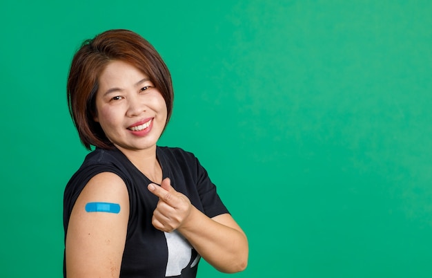 Foto foto de estudio de una paciente asiática de mediana edad sentada, sonrisa, mirada a la cámara, muestra de la mano del corazón mini y vendaje de yeso azul en el brazo después de recibir la vacuna contra el coronavirus covid 19 sobre fondo verde.