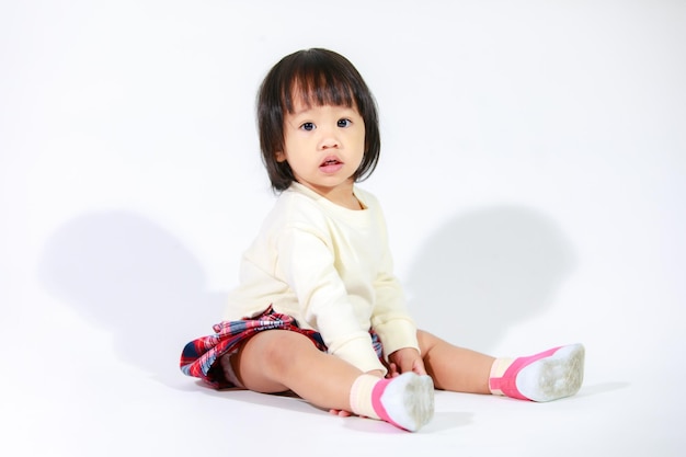 Una foto de estudio de una pequeña y linda modelo asiática de cabello negro corto con una falda a cuadros informal sentada y sonriendo sobre fondo blanco