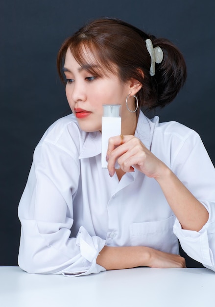 Foto de estudio de primer plano de una joven asiática glamurosa modelo femenina de moda con maquillaje en una camisa blanca casual sentada sonriendo mirando a la cámara sosteniendo una botella de crema facial sobre fondo negro