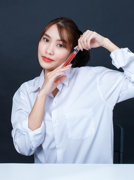 Foto de estudio de primer plano de retrato de modelo femenino de moda urbana de glamour joven asiático con maquillaje en camisa blanca casual sentado sonriendo mirando a la cámara sosteniendo lápiz labial rojo sobre fondo negro.
