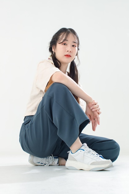 Foto de estudio de recorte aislado de retrato de modelo de moda adolescente linda joven asiática con peinado de trenzas de coletas en ropa casual de moda sentada en el suelo posando mirando a la cámara sobre fondo blanco