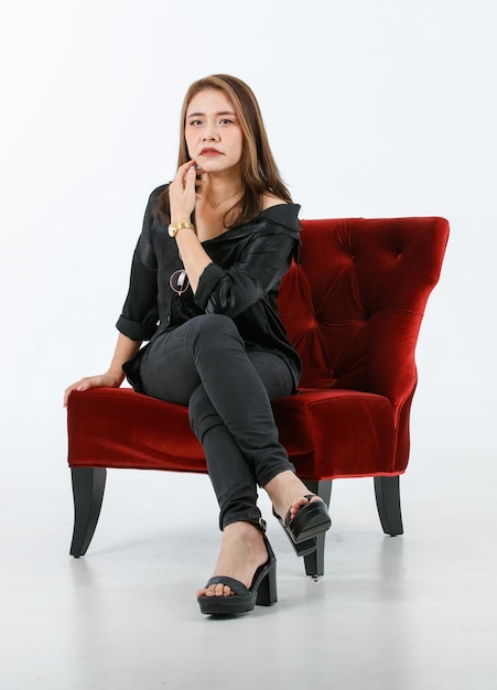 Foto de estudio de retrato de una alegre modelo asiática viajera de moda vestida con un traje negro informal, tacones altos, sentada sonriendo en un sillón rojo, mira la cámara con fondo blanco