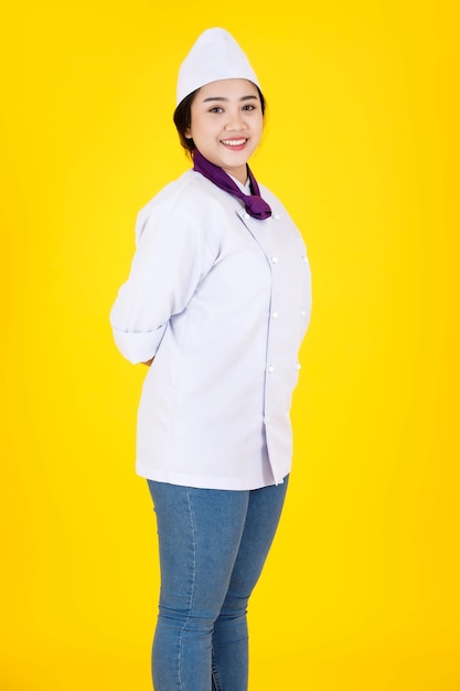 Foto de estudio de retrato de Asia profesional exitoso elegante confiado restaurante del hotel chef ejecutiva femenina en uniforme de cocina blanco de pie sonriendo mirar a la cámara con los brazos cruzados sobre fondo amarillo.