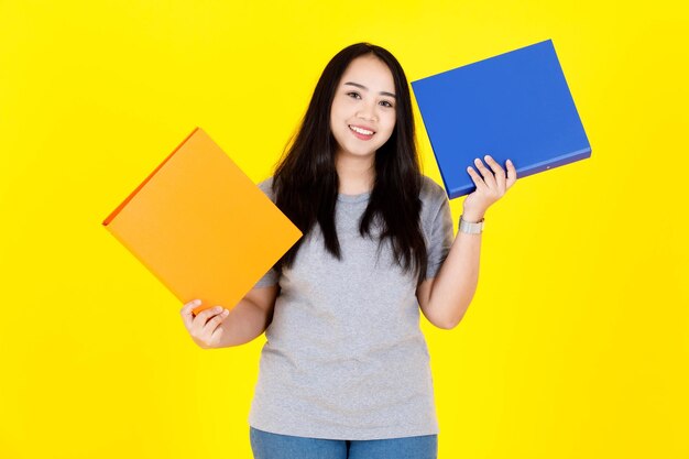 Foto de estudio de retrato de asiática feliz joven gordita regordeta pelo largo negro modelo estudiante en traje casual de pie sonriendo sosteniendo coloridas carpetas de archivos de documentos de papeleo sobre fondo amarillo.