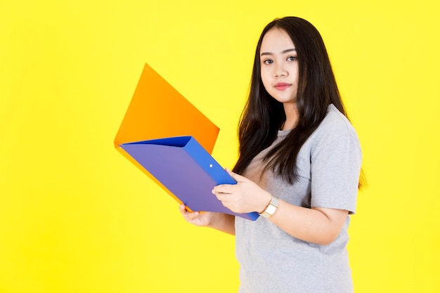 Foto de estudio de retrato de asiática feliz joven gordita regordeta pelo largo negro modelo estudiante en traje casual de pie sonriendo sosteniendo coloridas carpetas de archivos de documentos de papeleo sobre fondo amarillo.