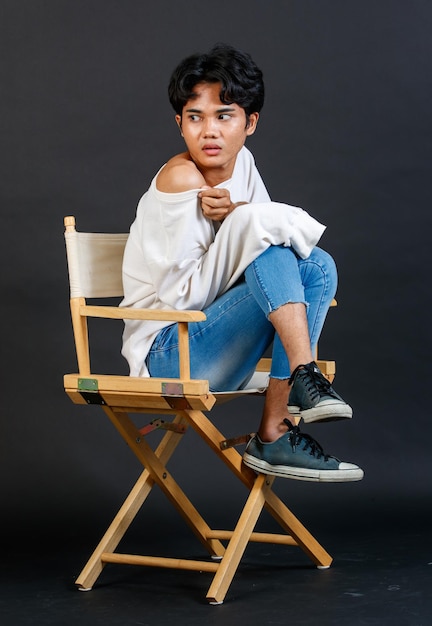 Foto de estudio de retrato de cuerpo completo del joven asiático LGBTQ gay glamour guapo bisexual homosexual modelo masculino en ropa informal sentado con las piernas cruzadas en una silla de madera posando gesticulando sobre fondo negro