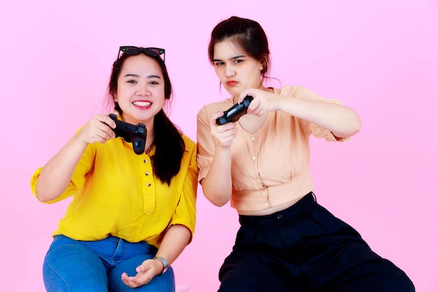 Foto de estudio de retrato de dos jóvenes asiáticos alegres competitivos amigas o hermanas gamer en ropa casual con gafas de sol sentado sonriendo sosteniendo joystick jugando juntos sobre fondo rosa.