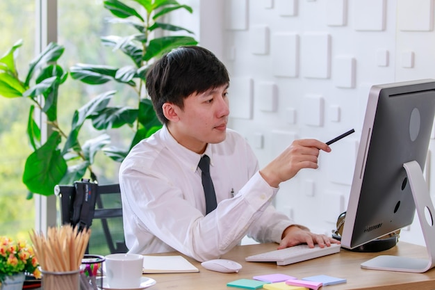 Foto de estudio de retrato de un exitoso hombre de negocios profesional asiático empleado en camisa formal con corbata sentado mirando la cámara en el escritorio de trabajo con monitor de computadora, teclado, mouse y papelería