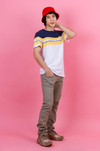 Foto de estudio de retrato de un joven y apuesto adolescente urbano asiático, modelo masculino de moda con ropa de estilo callejero, camiseta deportiva, pantalones beige y sombrero de cubo rojo, de pie, mirando a la cámara con fondo rosa