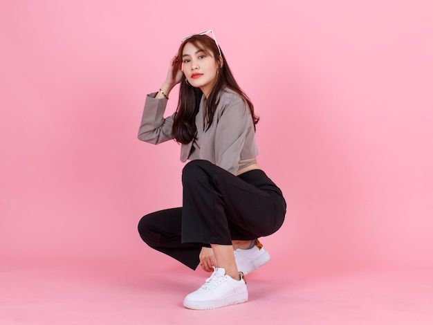 Foto de estudio de retrato de una joven asiática urbana de moda mujer hipster adolescente modelo de moda en la calle casual crop top usa chaqueta con gafas de sol en la cabeza sentada mirando a la cámara sobre fondo rosa