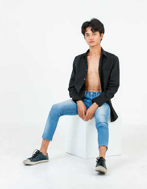 Foto de estudio de retrato de un joven asiático LGBT gay bisexual homosexual en topless modelo masculino de moda en camisa negra casual y jeans de mezclilla sentados en cajas cuadradas posando mirando a la cámara sobre fondo blanco