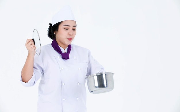 Foto de estudio de retrato de restaurante profesional asiático cocinando chef ejecutiva femenina en uniforme de cocinero y bufanda de pie sonriendo mirar a cámara sosteniendo una olla de acero inoxidable y tapa sobre fondo blanco.