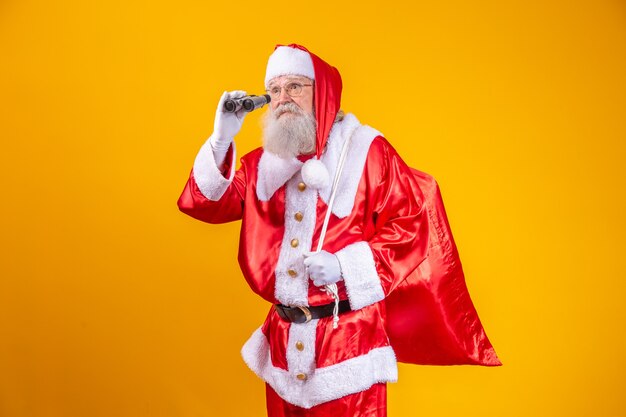 Foto de estudio de Santa Claus mirando a través de binoculares aislado sobre fondo amarillo