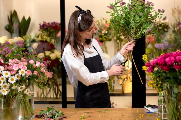 Foto foto de una exitosa floristería moderna que usa delantal creando un ramo de flores florecientes y coloridas en una moderna tienda floral interior, usando tijeras. concepto de negocio de pequeña tienda de flores